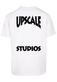 Biela bavlnená tričko s čiernym grafickým textom na zadnej strane: "UPSCALE" hore a "STUDIOS" dole v tučnom písme.