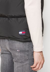 Schwarze gepolsterte Weste mit glänzender Oberfläche. Mit einem Tommy Jeans Logo-Patch an der Taille und einem hellen Langarmshirt darunter.