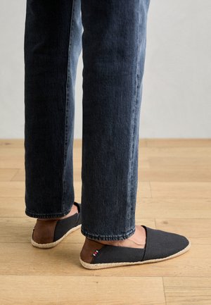 Chaussons en tissu gris avec une semelle tissée naturelle, ornés d'un petit accent tricolore. Portés avec un jean en denim sombre sur un sol en bois.