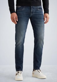 Slim-fit kavbojski jeans v temno modri barvi z zbledelimi detajli. Ima pet žepov in zadrgo. Ujemajo se z belimi in bež športnimi copati.