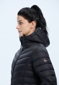 Mustekka puffer-takki korkealla kauluksella ja hupulla, jossa on vaakasuunnassa tikattuja osia ja logo-patch vasemmassa hihassa.