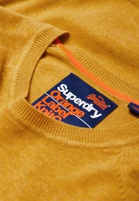 Maglione lavorato a maglia color senape con collo rotondo a coste, dotato di un'etichetta blu con la scritta "Superdry Orange Label" e dettagli di cucitura.