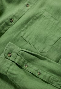 Camicia di lino verde con taschino sul petto, abbottonatura con bottoni scuri e una finitura di tessuto texturizzata.