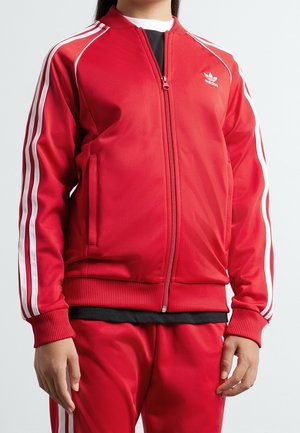 Personne portant un survêtement Adidas rouge avec des bandes blanches sur les manches et le pantalon, debout devant un fond clair uni.