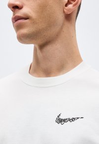 Κοντινό πλάνο στο κάτω μέρος του προσώπου και τον λαιμό ενός άνδρα που φοράει λευκό μπλουζάκι με στρογγυλή λαιμόκοψη και μικρό μαύρο αφηρημένο σχέδιο στο στήθος.