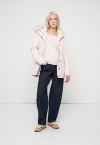 Ροζ παρκά με γούνινη κουκούλα, μπεζ πουλόβερ και σκούρο τζιν με λευκές τρουκς. Καφέ παπούτσια με διακοσμητικό μοτίβο.