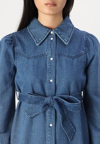 Denim haljina s ovratnikom, prednjim kopčanjem na dugmad i elastičnim rukavima. Ima pojas s vezanjem u struku i dva prednja džepa. Tamnoplava boja.