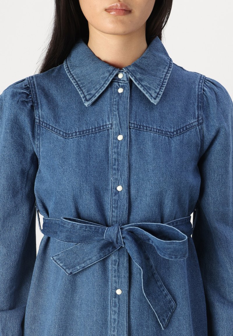Denim jurk met een kraag, knoopsluiting aan de voorkant en elastische mouwen. Voorzien van een gestrikte tailleband en twee voorzakken. Diepblauwe kleur.