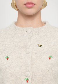 Cardigan tricotat de culoare gri deschis, cu guler rotund, având căpșuni roșii brodate și un accent cu broșă în formă de albină. Închidere cu butoane pe față.