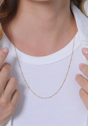 Collier fin en chaîne d'or avec de petits détails en perles porté autour du cou d'une personne, sur un t-shirt blanc côtelé.