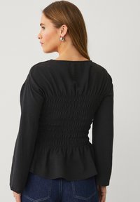 Zwarte blouse met lange mouwen; gerimpeld gedeelte bij de taille; textuurstof; ronde halslijn; peplum zoom; gedragen met denim onderkleding.
