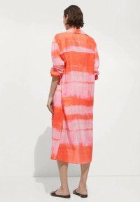 Langes, knopfbares Kleid im lebhaften Korallen- und Rosatöne-Tie-Dye-Muster, aus leichtem Stoff gefertigt, mit umgeschlagenen Ärmeln und seitlichen Schlitzen.