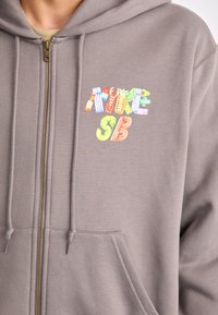Gray zip-up hoodie featuring an embroidered multicolored logo on the chest, with a fleece texture and drawstrings at the hood.