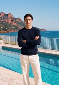 Jeune homme en pull marine et pantalon blanc debout près d'une piscine avec vue sur la montagne et l'océan sous un ciel bleu clair.