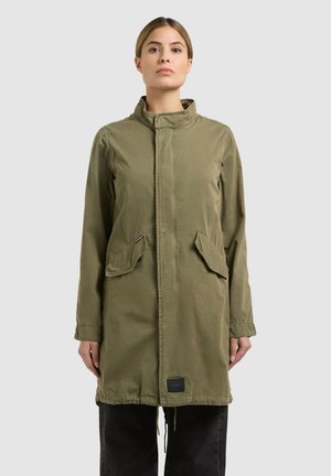 Femme portant une longue veste vert olive avec des poches à rabat, debout devant un fond uni gris clair.