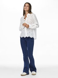 Kobieta uśmiechająca się, ubrana w białą bluzkę z koronki typu eyelet z falistym wykończeniem, ciemnoniebieskie dzwoniaste dżinsy i białe trampki, stojąca na białym tle.