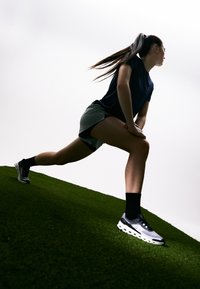 Sportschoenen in zwart, wit en grijs met een bovenwerk van gaas en een gezoneerde zool. Model draagt een marineblauw shirt en lichte shorts op groen gras.