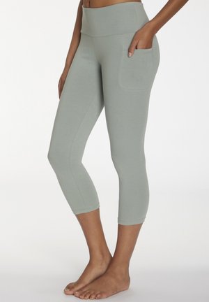 Woman wearing light grey cropped leggings with a high waistband and side pocket, standing barefoot against a plain background.