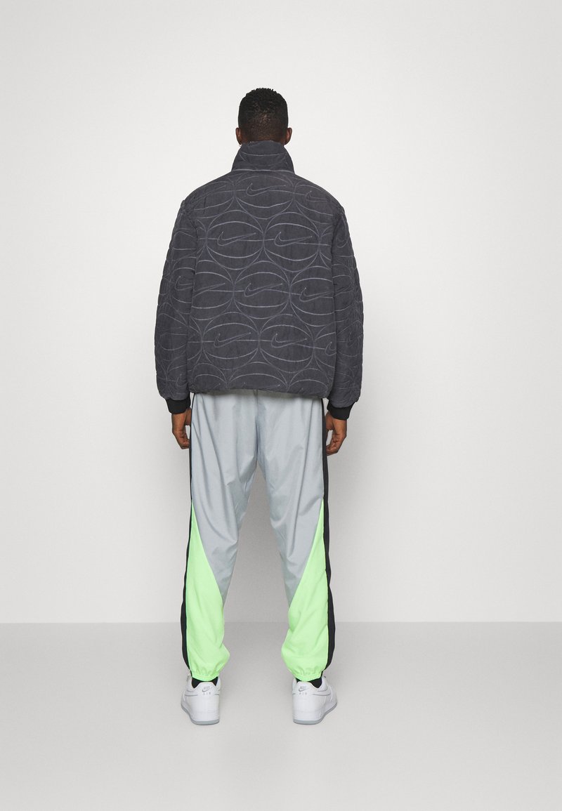 Giacca Nike grigia trapuntata con colletto alto, abbinata a pantaloni leggeri grigi con pannelli laterali verde neon. Scarpe bianche completano il look.