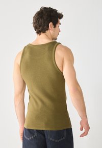 Débardeur côtelé vert olive avec une coupe ajustée, des emmanchures larges et une texture lisse. Porté avec un jean en denim foncé, vu de dos.