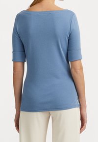 Donna con una maglietta blu a maniche corte con maniche a risvolto e pantaloni color crema, vista di spalle su uno sfondo semplice.