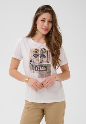 Ung kvinde med langt brunt hår bærer en hvid t-shirt med grafik og beige bukser, accessorized med guldbelagte øreringe, armbånd og ringe, smilende let.