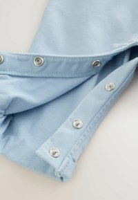 Tessuto azzurro chiaro con una texture liscia e bottoni a pressione argentati lungo il bordo, che mostrano un design con polsino piegato.