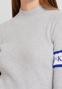 Maglione a coste grigio chiaro con collo alto. Presenta strisce blu e bianche con testo sulla manica. Texture morbida, design aderente.