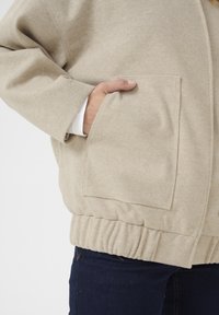 Veste en flanelle beige avec un tissu texturé, dotée de poches latérales et d'un ourlet élastiqué. Le design comprend une coupe décontractée et des coutures visibles.