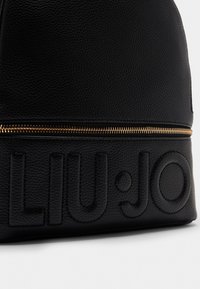 Schwarzer Leder-Rucksack mit strukturierten Oberflächen, goldener Reißverschlussbeschläge und eingraviertem Branding auf der Vorderseite. Kompakte, moderne Form.