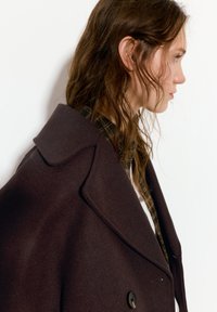 Tummanruskea villakangas takki, jossa on leveä kaulus, kaksirivinen malli ja hienovarainen tekstuuri. Napit ovat mustat ja pyöreät, mikä lisää yksityiskohtia etuosaan.