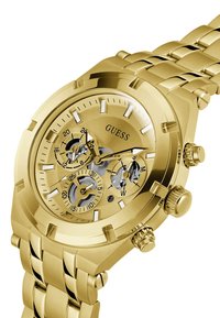 Montre analogique dorée avec une finition polie, présentant un cadran squelette argenté, trois sous-cadrans et un bracelet en métal maillé.