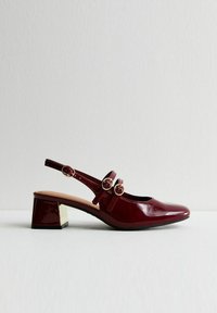 Burgunderfärgad lackskinnssandal med blockklack och tre guldspännen, visad från sidan på en vit yta.