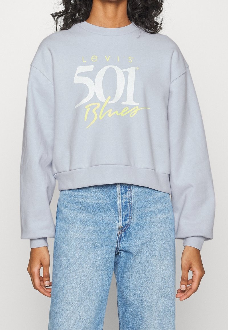 Personne portant un sweat Levi's 501 Blues gris clair et un jean taille haute bleu clair, debout devant un fond blanc.