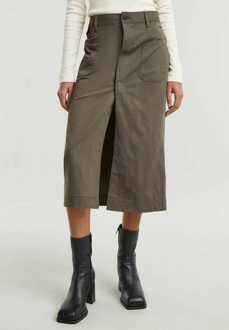 Gonna midi color khaki con spacco laterale, chiusura frontale con bottoni e due ampie tasche laterali. Abbinata a stivali neri in pelle con tacco.