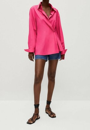 Vrouw draagt een oversized felroze gebuttonde blouse, blauwe denim shorts en zwarte platte sandalen met bandjes, staand tegen een effen achtergrond.