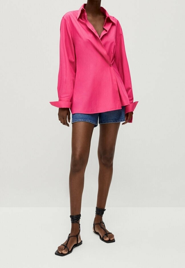 Femme portant une chemise rose vif oversize boutonnée, un short en denim bleu et des sandales plates noires à brides, debout devant un fond uni.