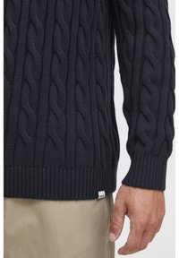 Marineblauer Kabelstrickpullover mit geripptem Saum, der dicke Zöpfe und ein kleines Stofflabel in der Nähe des Saums aufweist. Das Material wirkt weich und strukturiert.