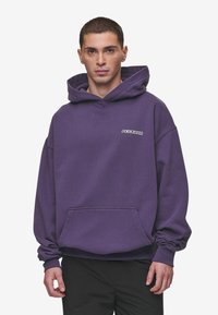 Sudadera con capucha de color púrpura, con un bolsillo frontal y un logotipo blanco de "PEGADOR", que presenta un corte holgado y una textura de tela suave.