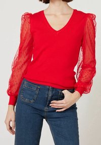 Pull rouge à col en V avec manches transparentes, bouffantes et à pois. Corps ajusté et poignets côtelés, associé à un jean en denim bleu.