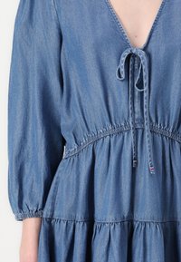 Džinsu kleita ar dziļu V veida kakla izgriezumu, saliektu jostasvietu un kupolveida piedurknēm. Iekļauj kakla saiti un volānu hemu vidēja zilā tonī.