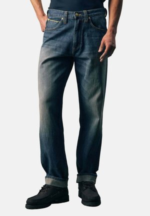 Mann trägt dunkelblaue, ausgewaschene Jeans mit aufgekrempelten Säumen und schwarze Schnürschuhe und steht vor einem hellen Hintergrund.
