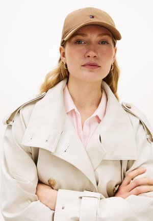 Beżowa czapka bejsbolowa z wyhaftowanym logo; beżowy trencz z guzikami i szerokimi klapami; pod spodem różowa koszula w kratę z kołnierzykiem.