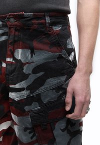 Κοντινό πλάνο ενός ατόμου που φοράει παντελόνι καμουφλάζ σε κόκκινο, γκρι και μαύρο χρώμα με μαύρο ριμπ μπλουζάκι, δείχνοντας το δεξί χέρι και τα δαχτυλίδια.