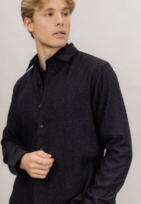 Mørkegrå, langermet skjorte i et mykt, teksturert stoff med knappestolpe foran og lett krage. Har fem knapper nedover placket.