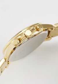 Montre en ton doré avec un boîtier rond, trois boutons-poussoirs et une couronne texturée, accompagnée d'un bracelet en métal poli et d'un fond minimaliste.