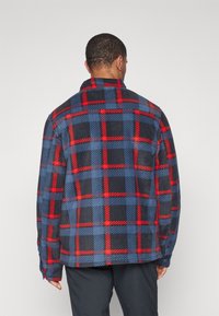 Modrá a červená kockovaná flanelová košeľa s dlhými rukávmi a stojatým golierom. Má veľký vzor a voľný strih. Mäkká textura.