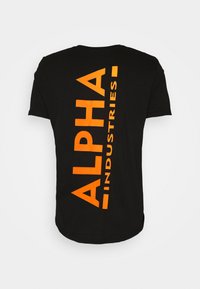 Camiseta negra con un gran texto naranja que dice "ALPHA INDUSTRIES" alineado verticalmente en la parte posterior. Material de algodón suave, cuello redondo.