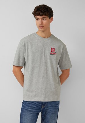 Junger Mann in grauem T-Shirt mit Harvard University-Logo und blauen Jeans, steht mit den Händen hinter dem Rücken und blickt zur Seite nach unten.