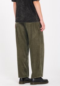 Pantaloni in corduroy verde con gamba larga, caratterizzati da righe verticali e una vestibilità rilassata. Scarpe nere con cuciture gialle completano l'outfit.